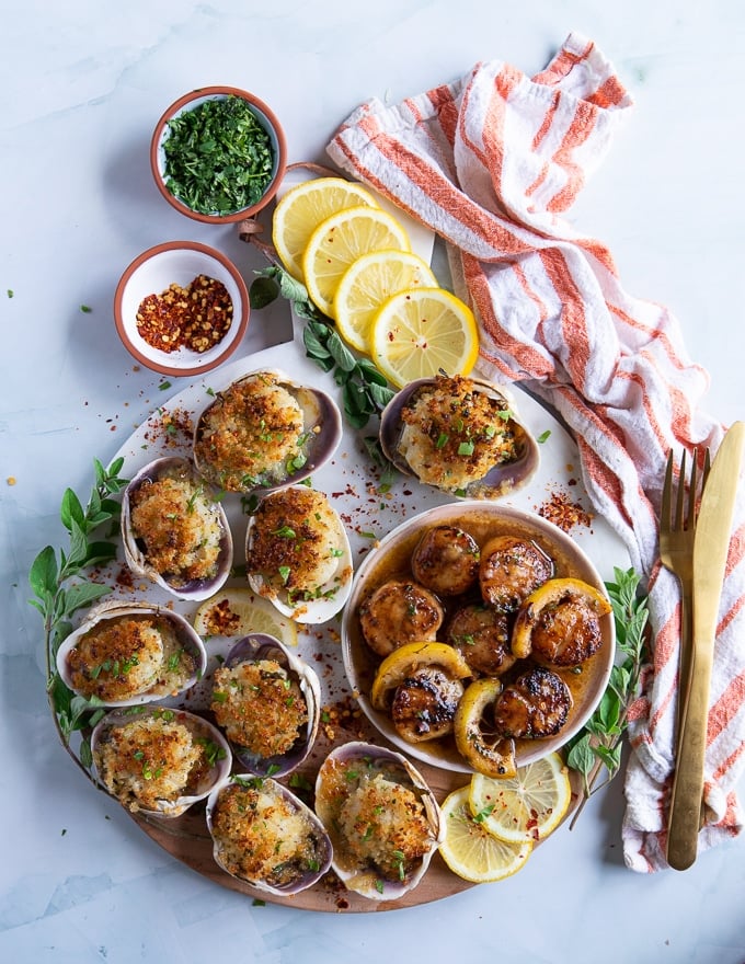 A board with two scallops recipes, a plate of seared scallops and a plate of baked scallops surrounded by lemon slices and a tea towel showing the different ways of how to cook scallops