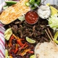 sheet pan fajitas with meat, veggies, tortillas, and all the toppings including cheese, sour cream, lettuce, avocado, salsa, lime wedges, sour cream