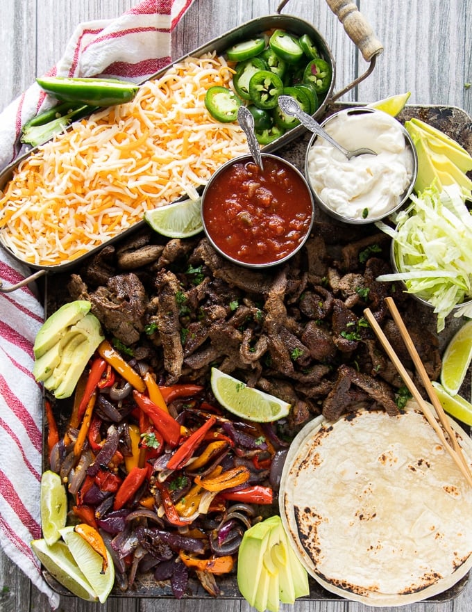 sheet pan fajitas with meat, veggies, tortillas, and all the toppings including cheese, sour cream, lettuce, avocado, salsa, lime wedges, sour cream
