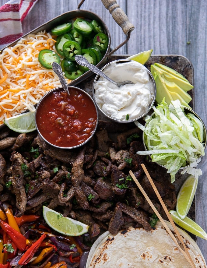 close up of the meat in sheet pan fajitas with a bowl of salsa and sour cream near by and lime wedges 