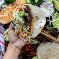 a hand holding an assembled fajita with tortilla, cooked veggies and meat, some jalapenos, cheese, salsa, avocado, sour cream