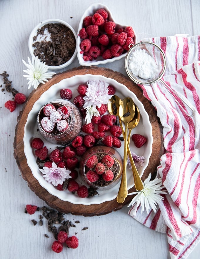 Plate with chocolate pudding in clear glass dishes with raspberries on top and gold spoons.