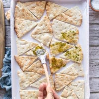 Pita chips arranged on a baking sheet and a hand brushing the olive oil and spice mixture on the pita chips