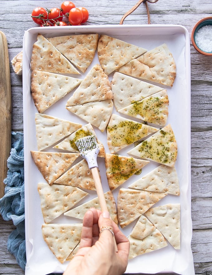 Pita chips arranged on a baking sheet and a hand brushing the olive oil and spice mixture on the pita chips