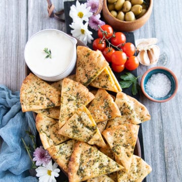 A plate of pita chips on a wooden plate surrounded by a whipped feta dip and some tomatoes, and coarse sea salt