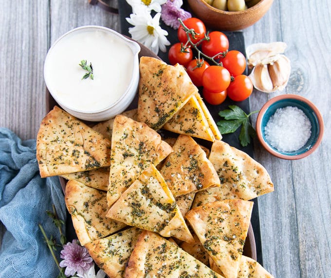 A plate of pita chips on a wooden plate surrounded by a whipped feta dip and some tomatoes, and coarse sea salt