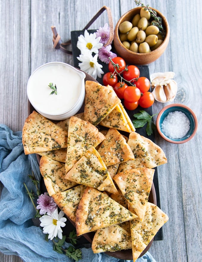 A plate of pita chips on a wooden plate surrounded by a whipped feta dip and some tomatoes, and coarse sea salt