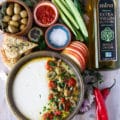 Top view of a plate of whipped feta surrounded by olives , cucumbers, hot sauce and pita chips