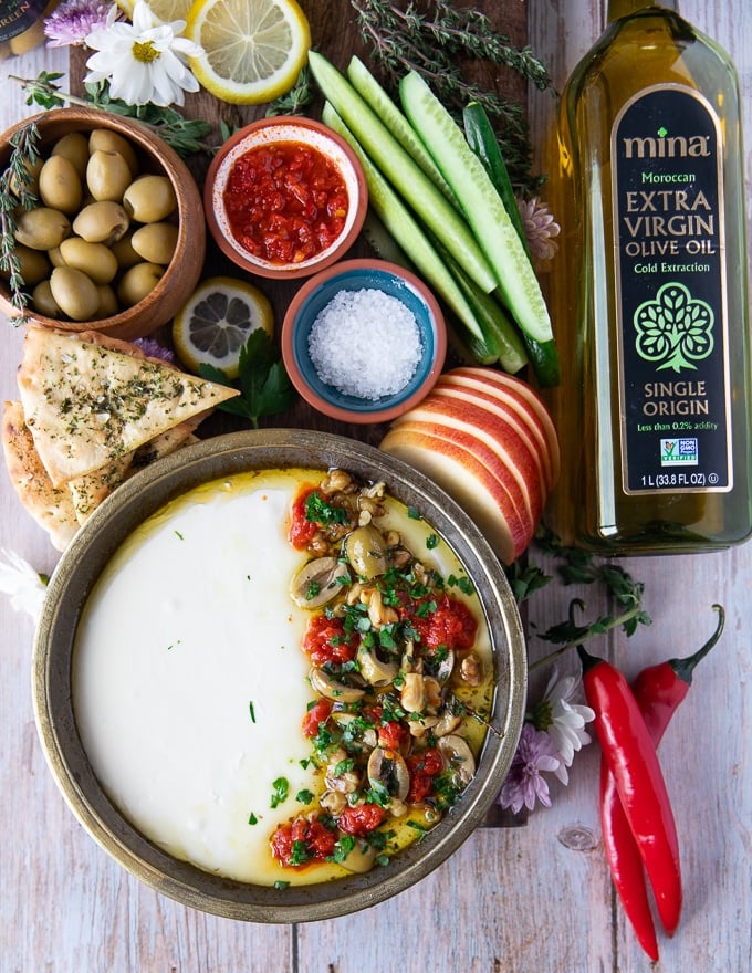 Top view of a plate of whipped feta surrounded by olives , cucumbers, hot sauce and pita chips