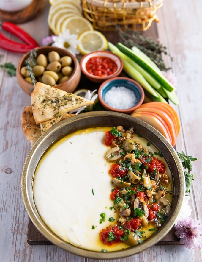 A whole board of whipped feta dip on a plate, some pita chips, cucumbers, apples, olives, Moroccan olive oil, some fresh herbs