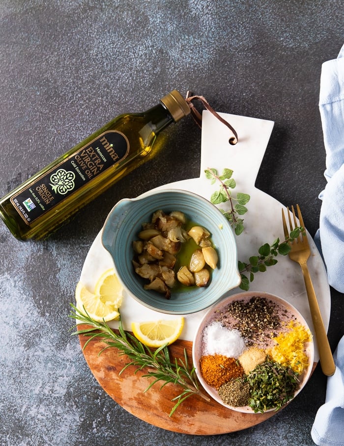 ingredients for air fryer whole chicken including a bow of roast garlic, a plate with different spices, fresh rosemary and lemon zest, and a bottle of olive oil