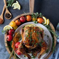 whole chicken from the air fryer rotisserie style on a plate surrounded by tomatoes, lemon slices, fresh rosemary and a fork and knife to cut