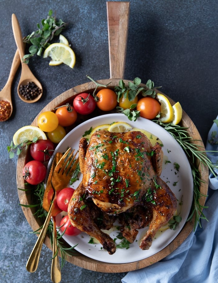 whole chicken from the air fryer rotisserie style on a plate surrounded by tomatoes, lemon slices, fresh rosemary and a fork and knife to cut