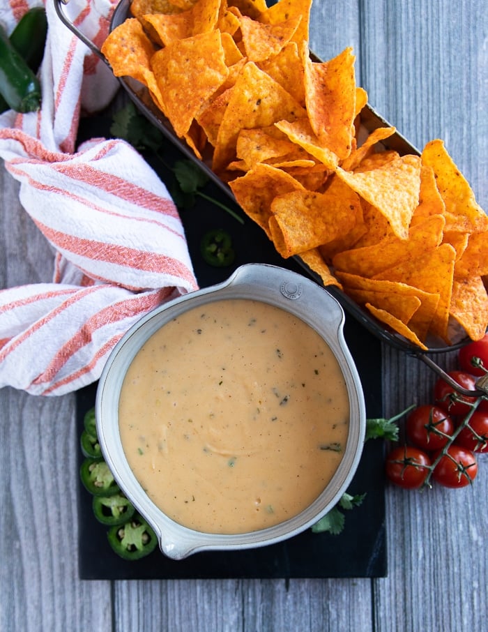Nacho cheese sauce poured into a bowl served along side chips, some tomatoes and a tea towel