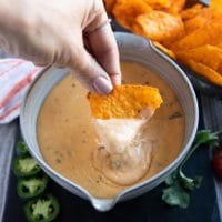 A hand going in to the bowl of nacho cheese sauce with a tortiall chips and showing the stretchy cheesy smooth sauce