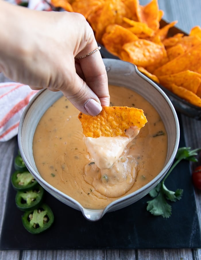 A hand going in to the bowl of nacho cheese sauce with a tortiall chips and showing the stretchy cheesy smooth sauce