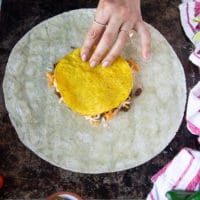 A tostada being put over the shredded cheese in the middle of the large tortilla building up the wraps