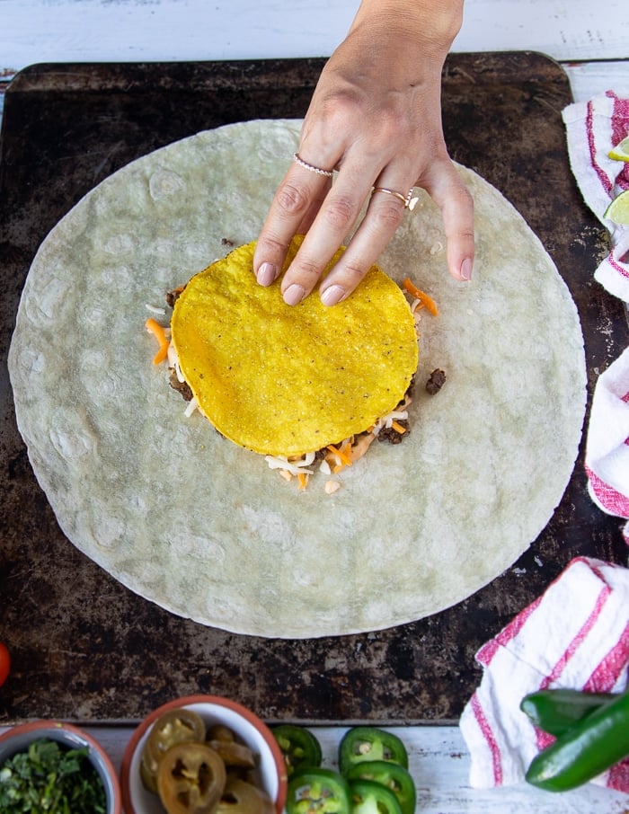 A tostada being put over the shredded cheese in the middle of the large tortilla building up the wraps