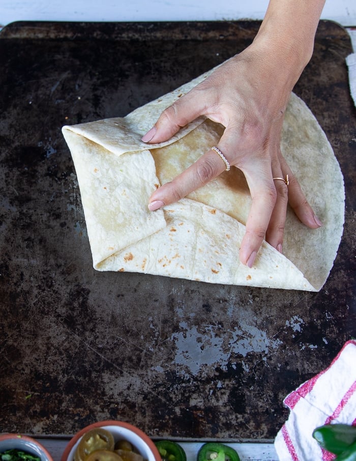 continuing the folding process with one hand holding down both sides and the the other hand folding the third side of the tortilla