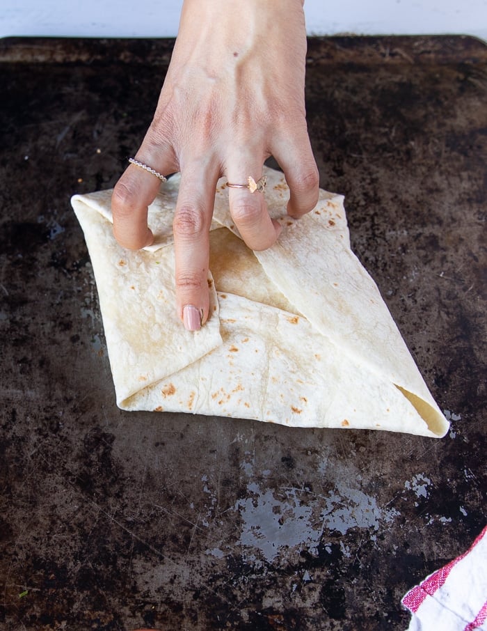 continuing the folding process of the curnchwrap supreme with one hand holding the pleats down while folding over the final side of the tortilla