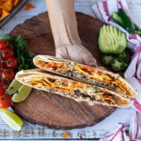 A hand holding one crunchwrap supreme cut in half showing the layers of seasoned meat, lettuce, cheese, sour cream, tomatoes, cheese sauce, avocado and tortilla