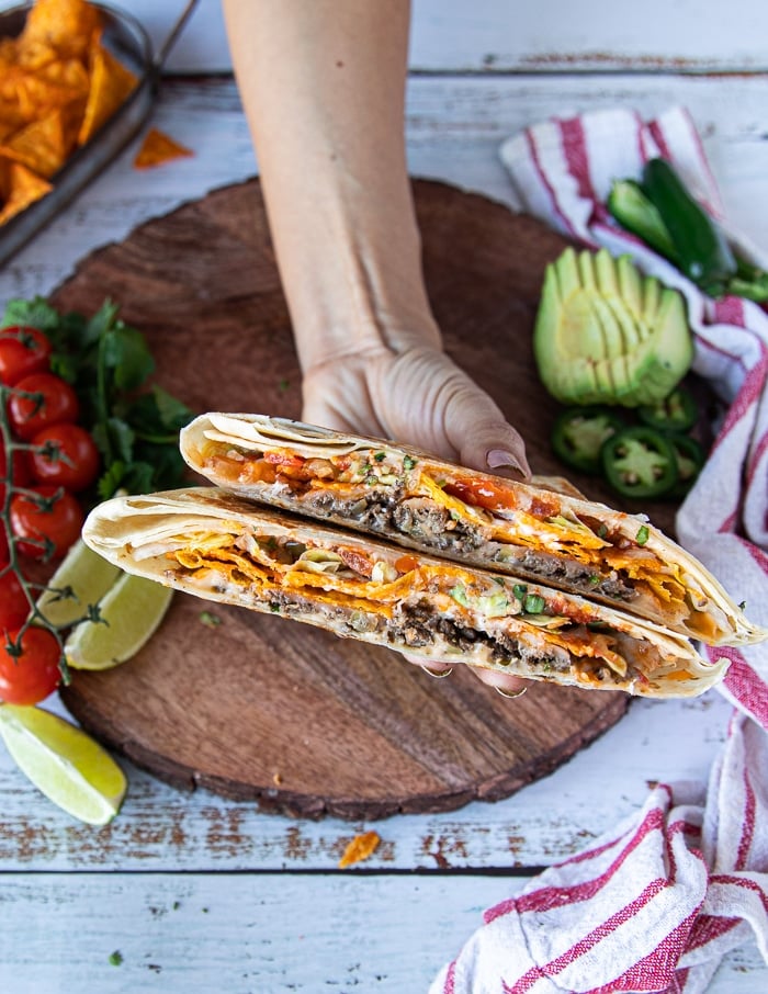 A hand holding one crunchwrap supreme cut in half showing the layers of seasoned meat, lettuce, cheese, sour cream, tomatoes, cheese sauce, avocado and tortilla