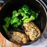 chicken breasts in an air fryer basket fliped half way and ready to continue cooking. A hand adding in some broccoli next to the chicken breasts to air fry them with the chicken to make a full meal