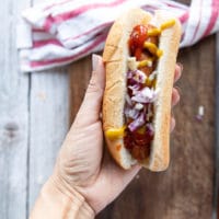 a hand holding an air fryer hot dog placed in toasted bun and dressing it with ketchup, onions, relish and mustard