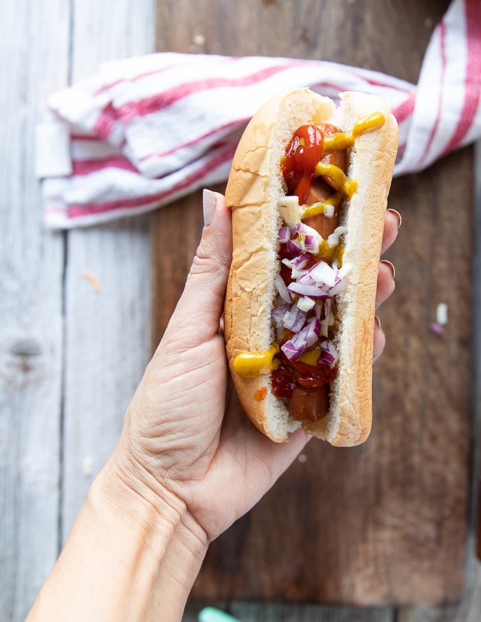 a hand holding an air fryer hot dog placed in toasted bun and dressing it with ketchup, onions, relish and mustard