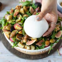 a hand holding a ball of burrata cheese ready to place it overt the salad