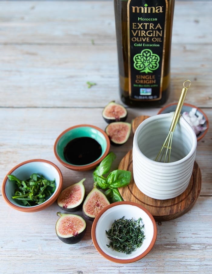 a small pouring jar next a small bowls of salad dressing ingredients including olive oil, shallots, fresh herbs, balsamic syrup and salt