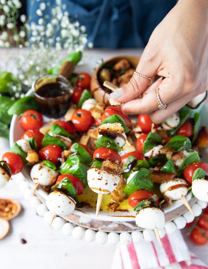 a hand sprinkling some croutons over the caprese skewers