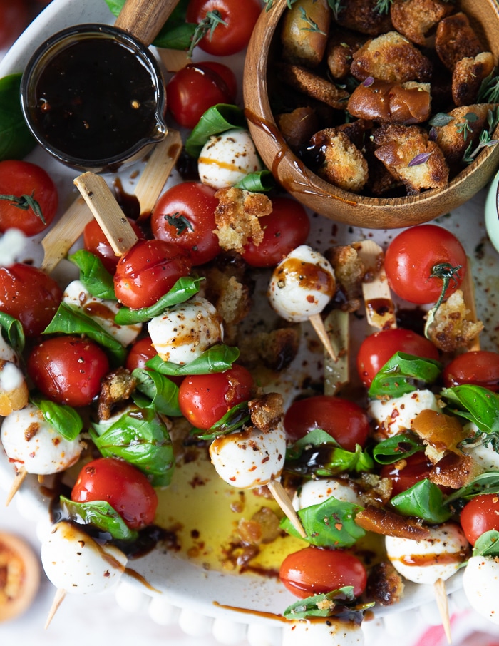close up of a skewer of caprese salad over olive oil and balsamic glaze, seasoned with chilli flakes and a bowl of croutons