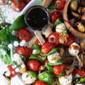 A Caprese skewer on a plate with a bowl of balsamic glaze on the side and fresh basil leaves