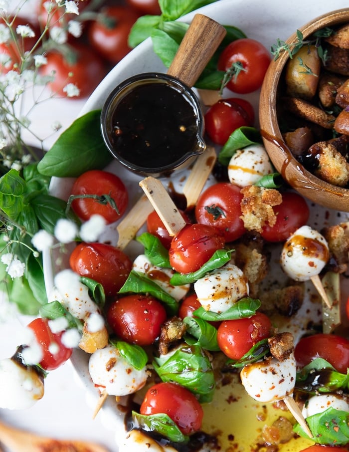 A Caprese skewer on a plate with a bowl of balsamic glaze on the side and fresh basil leaves