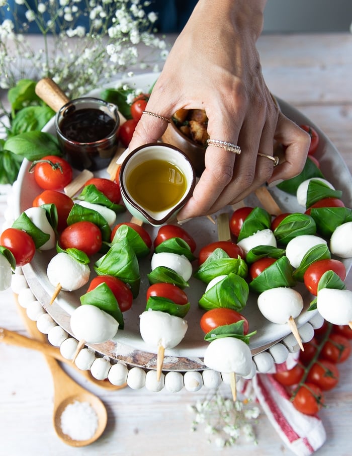 A small jar with olive oil being poured over the caprese skewers