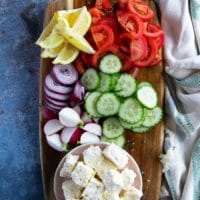 A wooden board of sliced cucumbers, tomatoes, feta, onions, lemon wedges, radishes