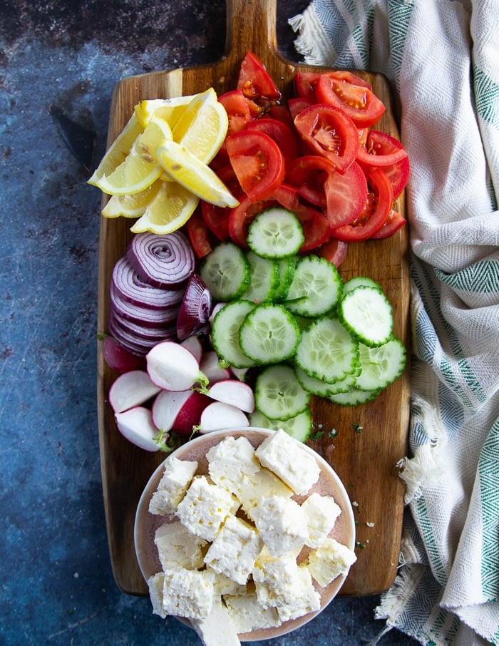 A wooden board of sliced cucumbers, tomatoes, feta, onions, lemon wedges, radishes