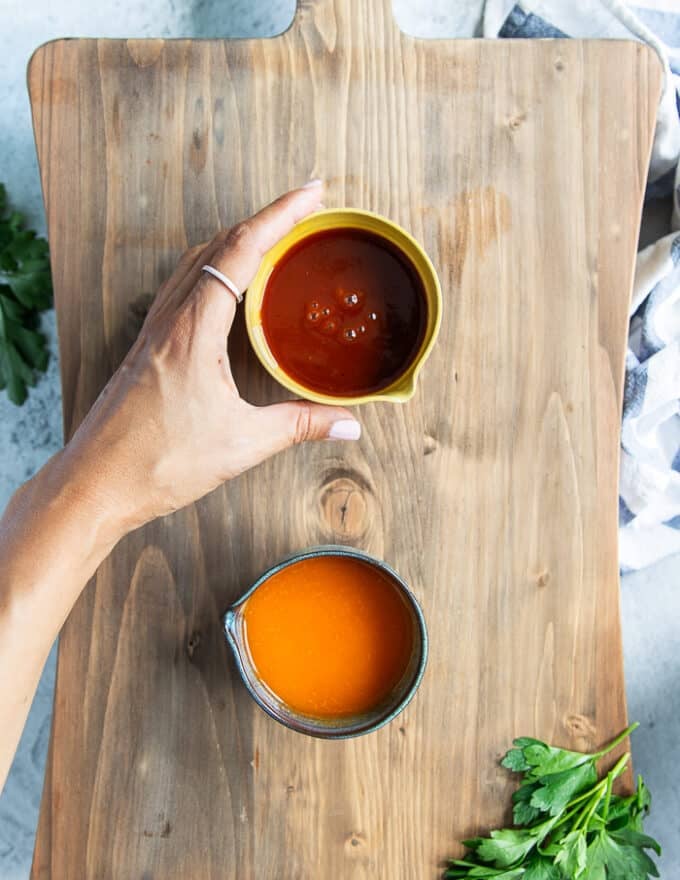 A hand holding two sauces to toss into the chicken wings, one hot wing sauce and one bbq sauce