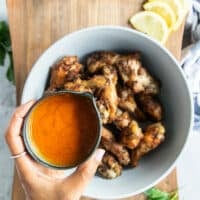 A hand pouring the hot sauce over the grilled chicken wings in a bowl.