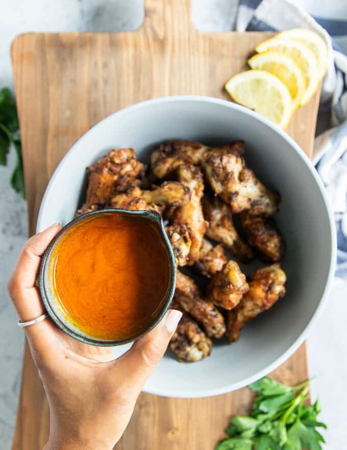 A hand pouring the hot sauce over the grilled chicken wings in a bowl.