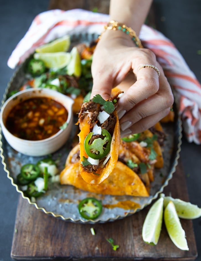 A hand holding a lamb birria over a plate of birria tacos