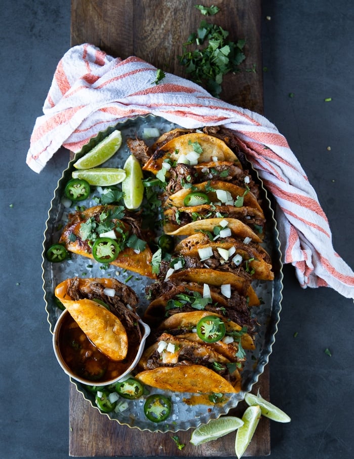 a plate of lamb birria tacos with the birria dipping sauce over a wooden board and some lime wedges