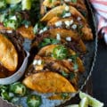 A row of lamb birria tacos arranged on a metal plate sprinkled with cilantro, onions and jalapeños