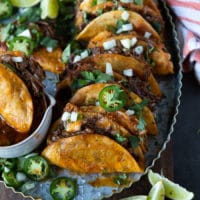 A row of lamb birria tacos arranged on a metal plate sprinkled with cilantro, onions and jalapeños