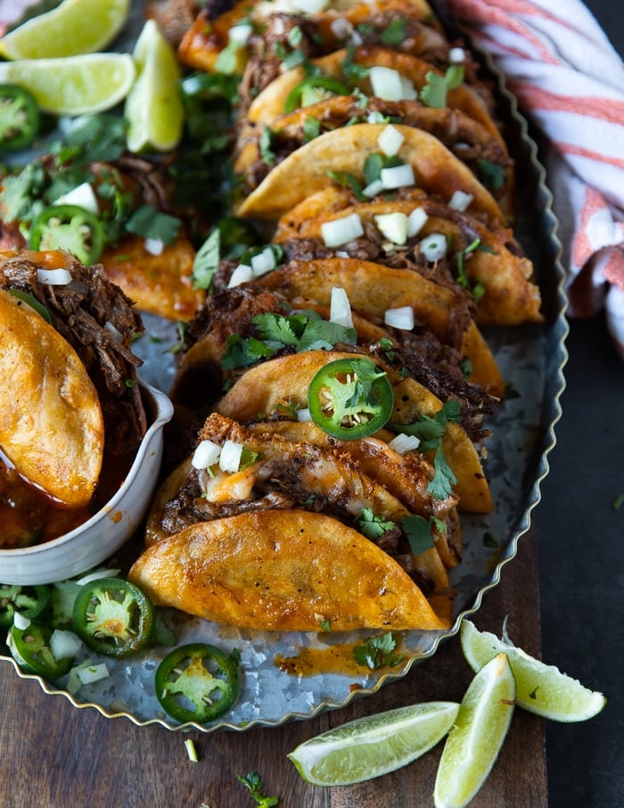 A row of lamb birria tacos arranged on a metal plate sprinkled with cilantro, onions and jalapeños 