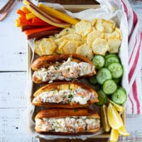 A tray of lobster roll sandwiches, some chips on the side, cucumbers, carrots and a kitchen towel. The lobster roll is stacked side by side
