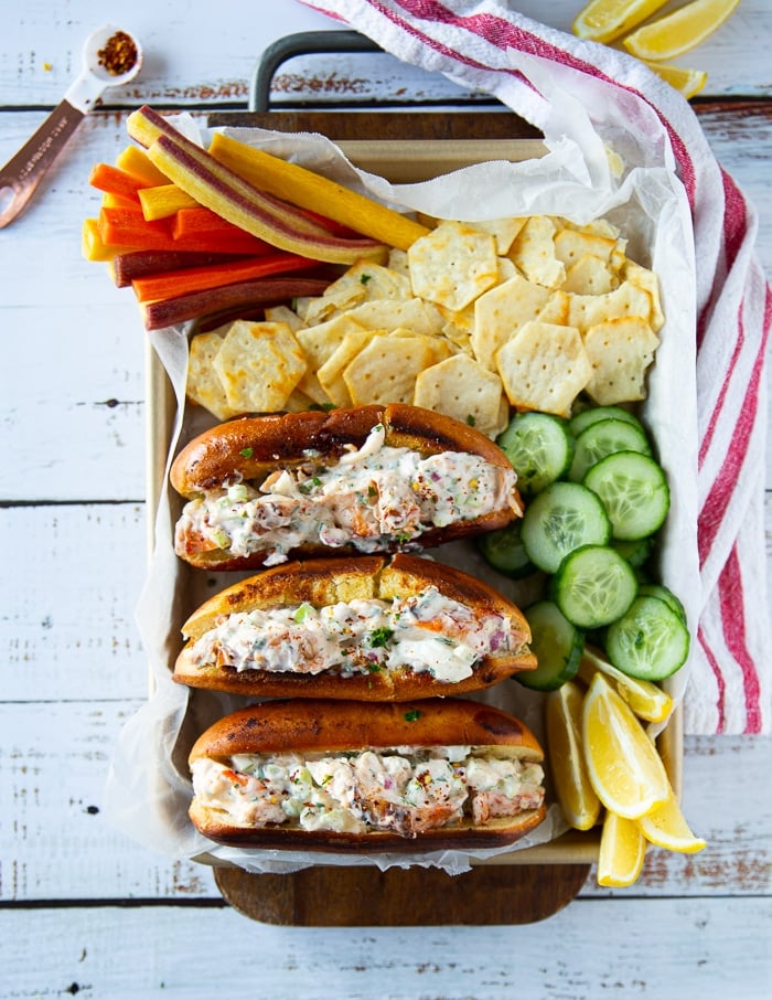 A tray of lobster roll sandwiches, some chips on the side, cucumbers, carrots and a kitchen towel. The lobster roll is stacked side by side