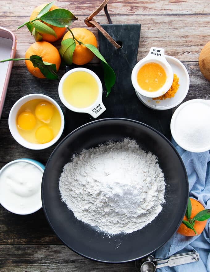 Ingredients for mandarin orange cake recipe including a bowl of flour, a bowl of sugar, a bowl of eggs, a bowl of oil, mandarin orange juice, zest of mandarin orange, baking soda