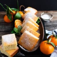 A plate of cut up mandarin orange cake squares dusted with powdered sugar and surrounded by mandarin oranges